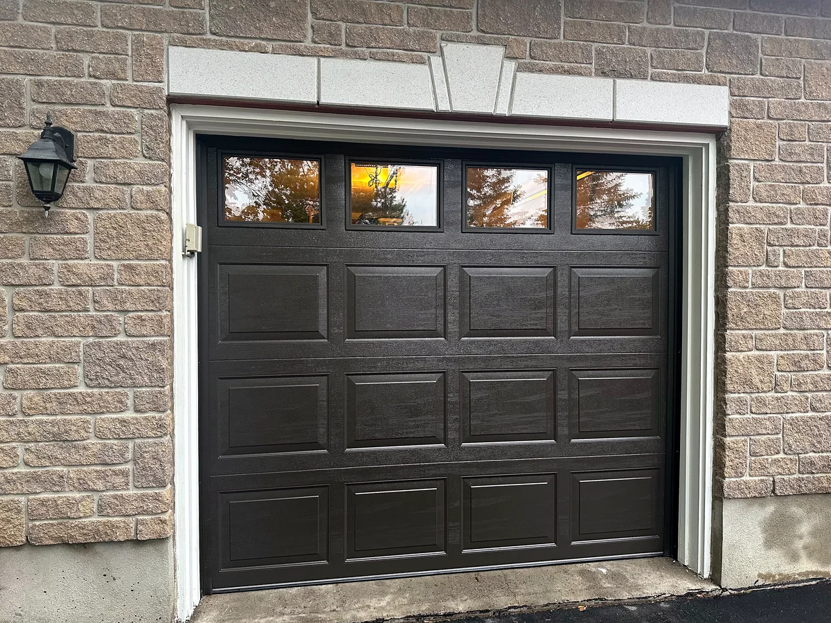 Black raised-panel garage door with four top windows in a brick opening