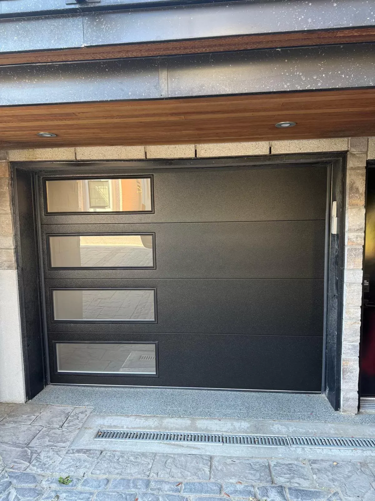 Modern black flush garage door with four offset windows down the left side on a Calgary stone garage
